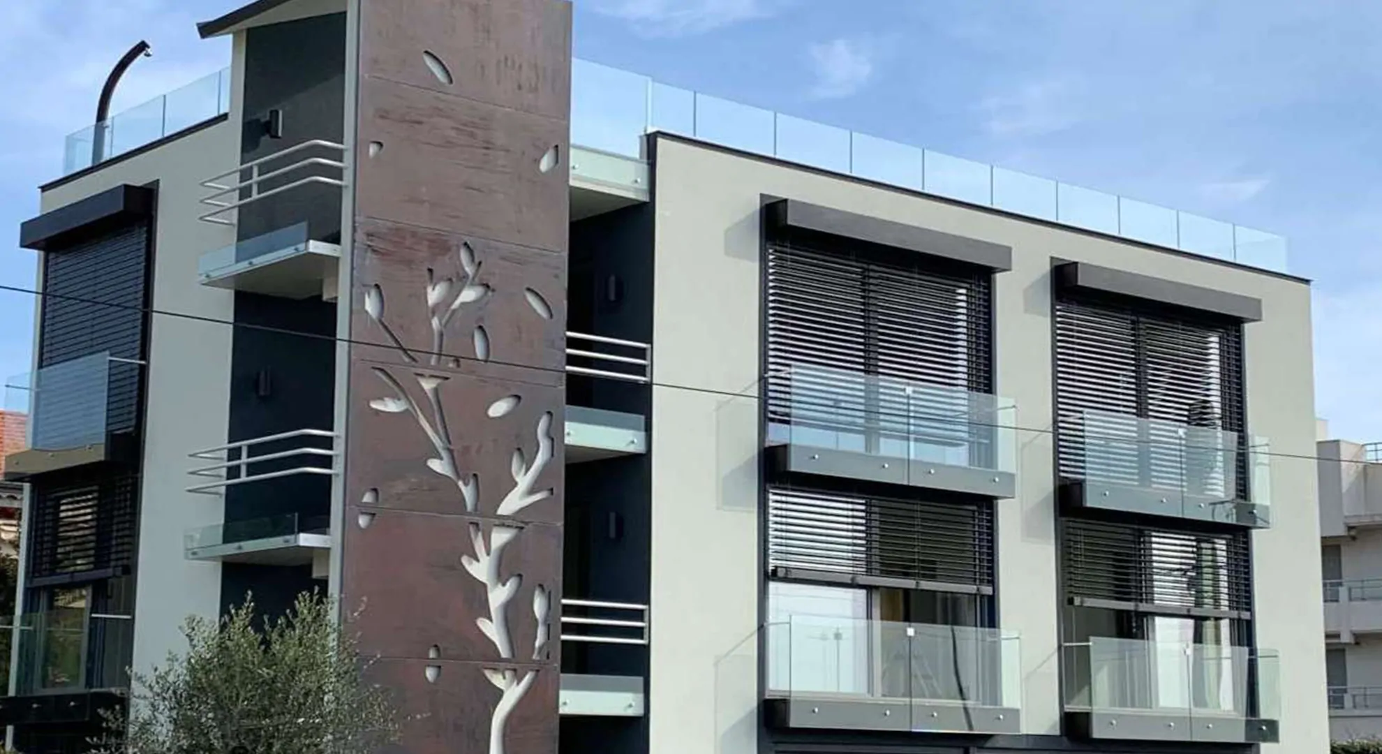 La facciata moderna di questo edificio residenziale gioca con le trasparenze delle ampie vetrate, parapetti in vetro trasparente ed il pannello decorativo con motivi vegetali, dinamico dialogo tra luce ed architettura.